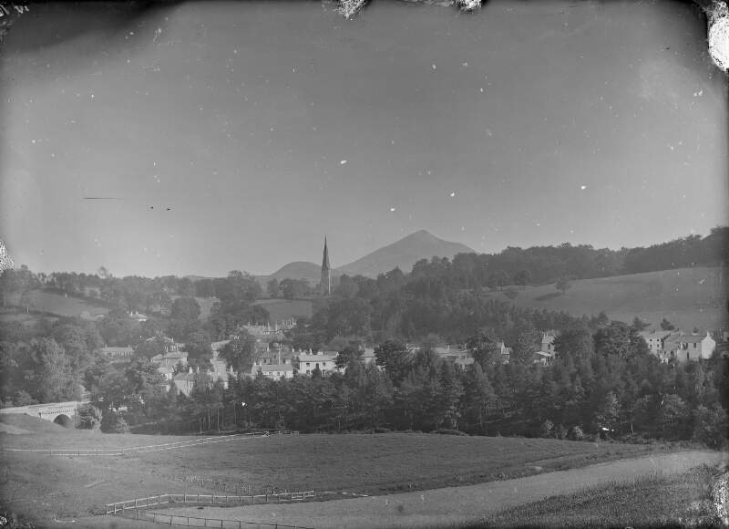[Town or village with church tower visible in the distance surrounded by trees and grass land.]