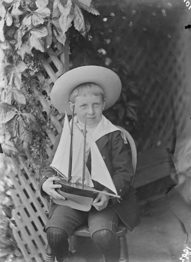 [Young boy in a sailor suit holding a toy sailing boat on his lap in front of trellis.]