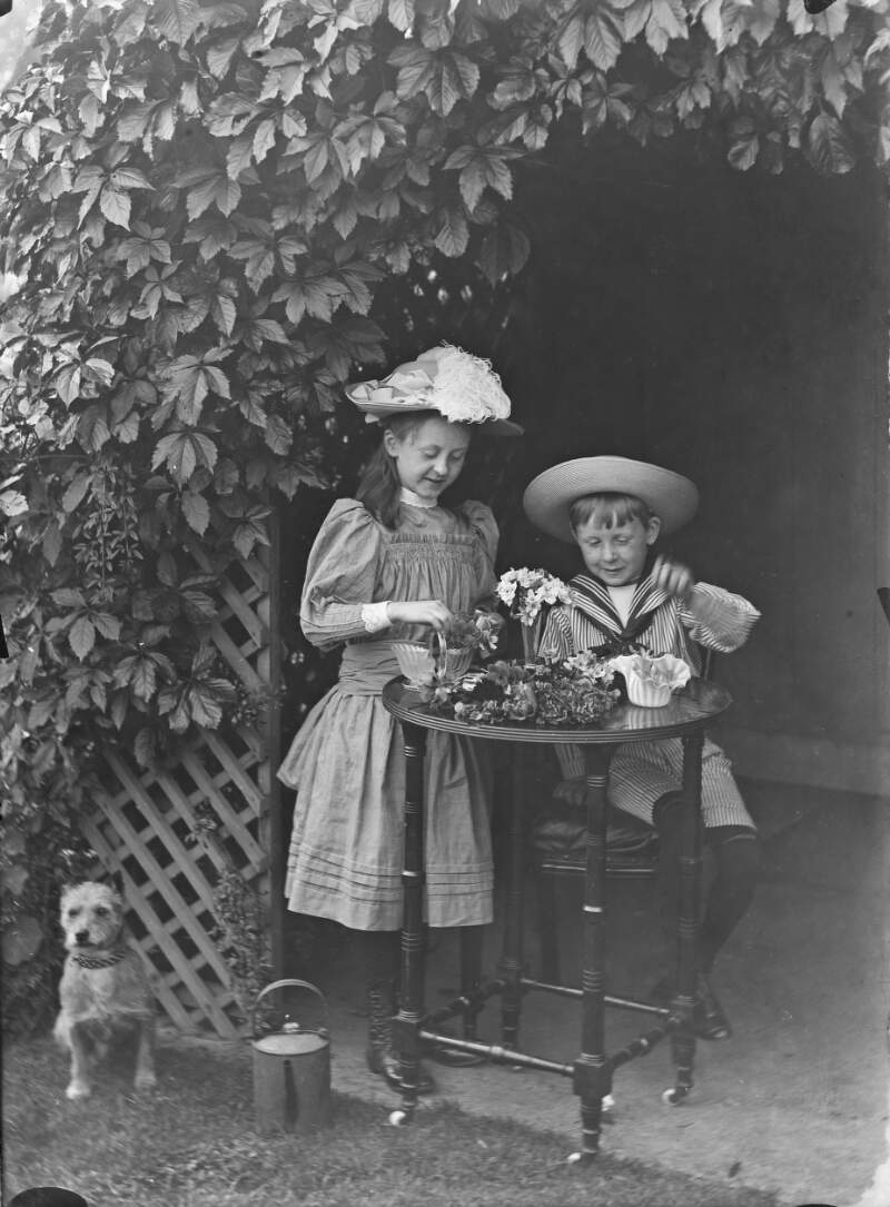 [A boy dressed in a sailor suit and an older girl playing with flowers on a small round table near a wooden trellis.]