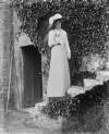 [Woman with parasol dressed in white standing on stone steps near an ivy-covered wall with an entrance.]