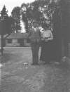 [A young man with an older woman standing by a tree with stables in the background.]