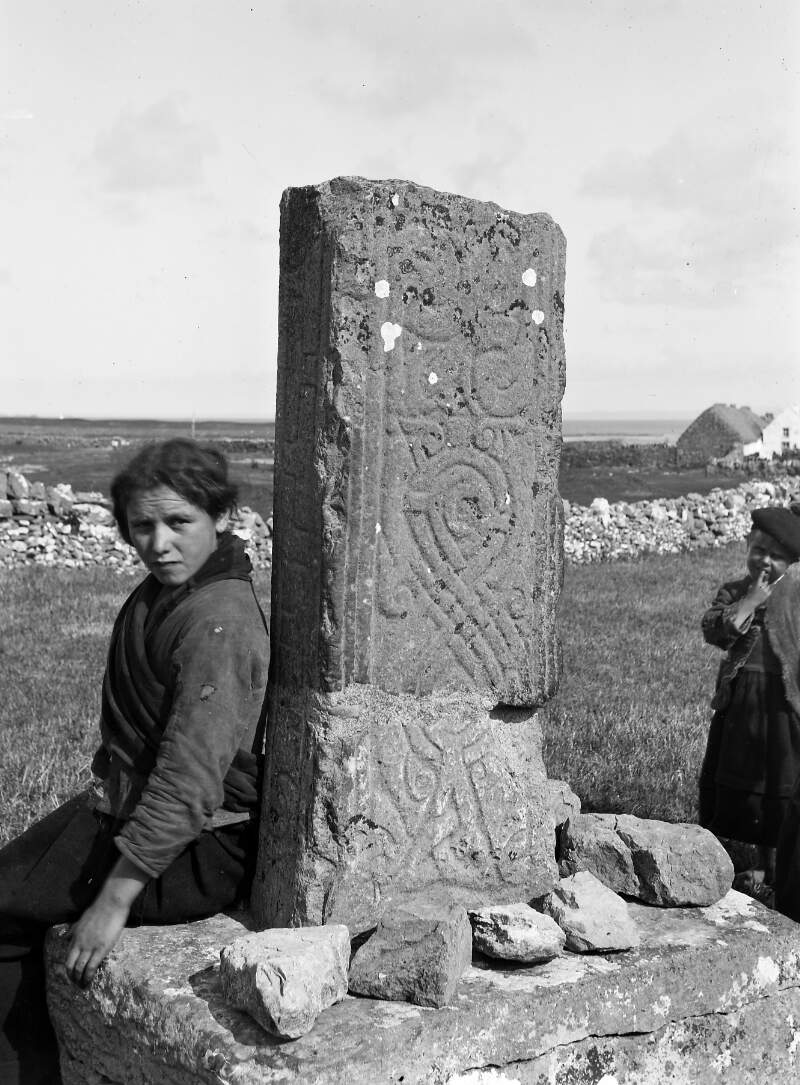 [Woman seated next to the west face of the shaft of a broken High Cross at Cill Éinne, Inis Mór, Aran Islands, Co. Galway. A child looks on in the background.]