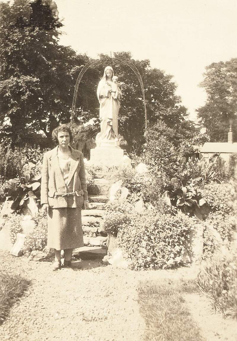 [Woman standing beside statue of the Virgin Mary in an unidentified garden]