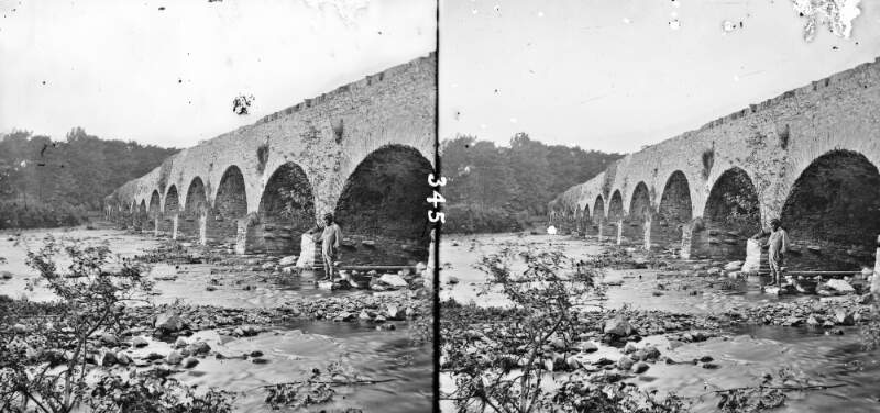 Bridge of possibly 15 arches, possibly over river Laune, seen from river bed or bank, man in foreground, with stick/rod, Killarney, Co. Kerry
