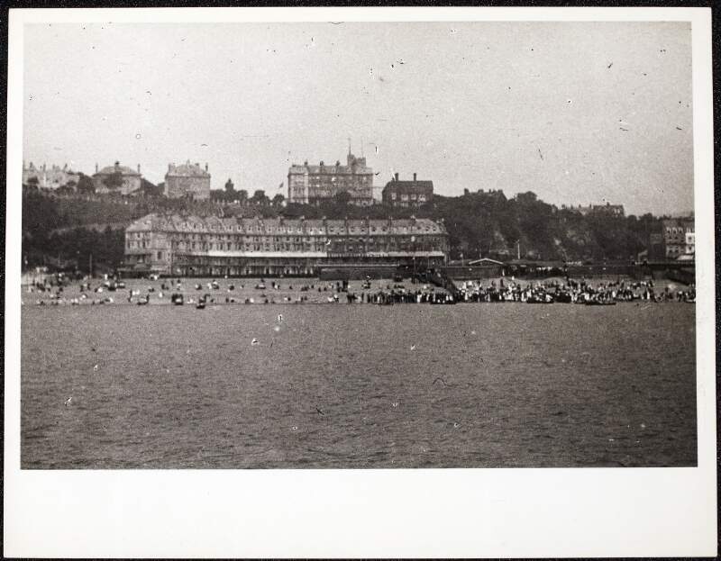 [Crowd on beach with row boats, with crescent of buildings at ground level and other large buildings on headland, taken from sea, location unidentified]