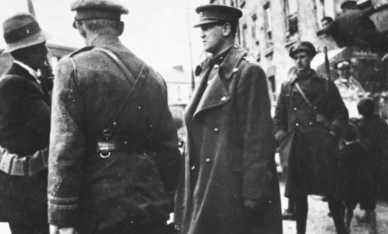 [Michael Collins with unidentified soldiers, gathered on an unidentified street with an armoured car in background]
