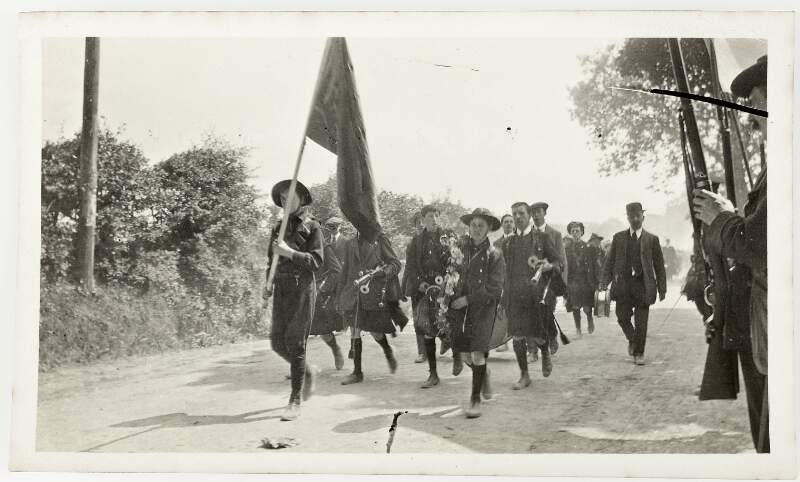 [Unidentified children and men marching along country road carrying instruments and a flag, including men lined along roadside holding guns]