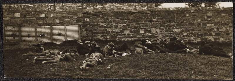 [Unidentified men laying low on grass verge alongside a stone wall, with some men in uniform carrying guns]