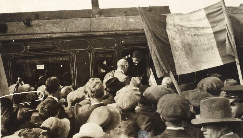 [Éamon De Valera disembarking a train to an awaiting crowd, including a tri-colour flag]