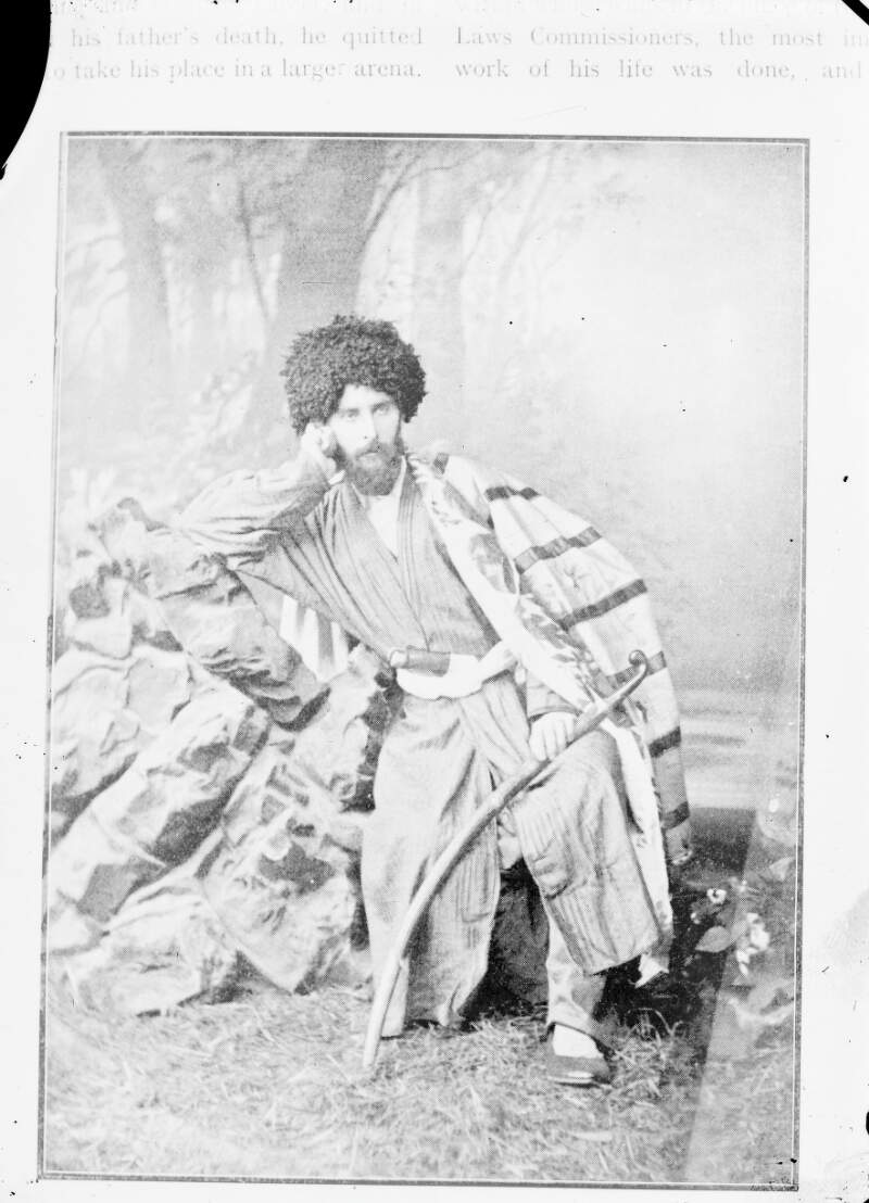 [Unidentified man in Asian dress with sabre, and draped in unknown flag, leaning against rock, full-length portrait]