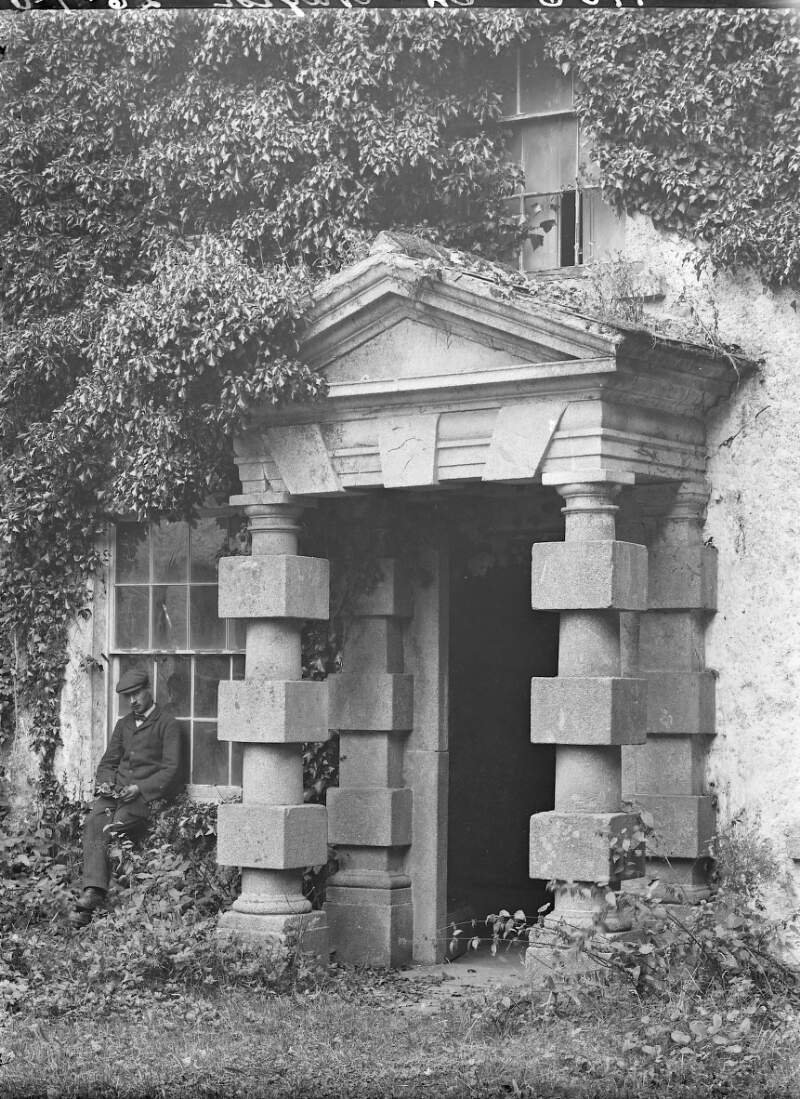 [Stone portico of unidentified building, overgrown with ivy and foliage, with man in suit and flatcap sitting on window sill]