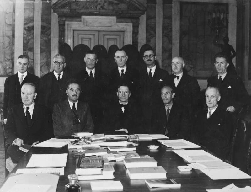 [Éamon De Valera seated front and centre at a large table covered in papers and books, surrounded by members of the governement, including Frank Gallagher]