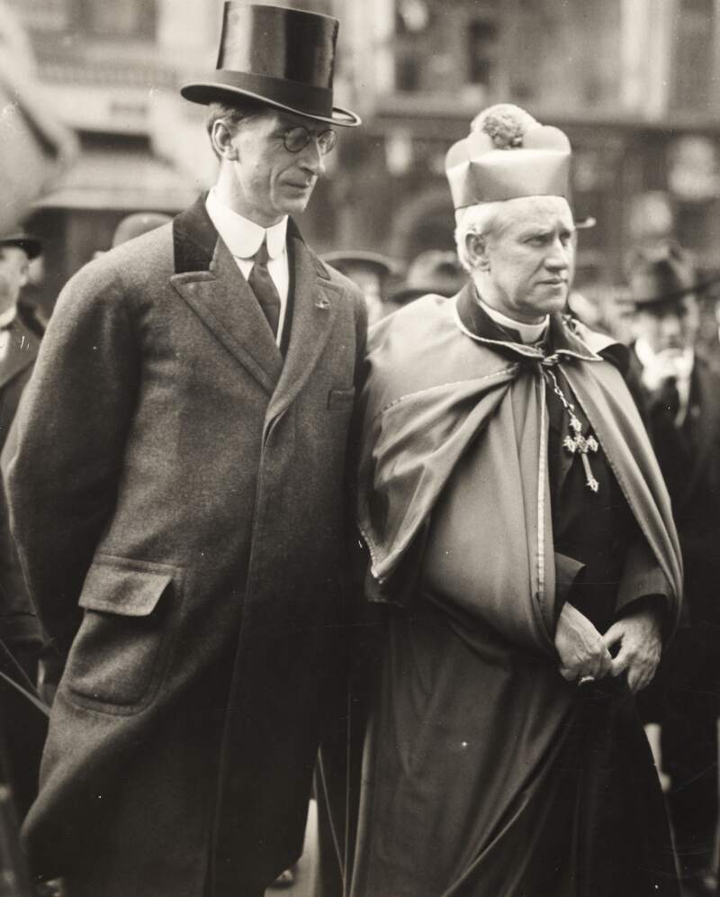 [Éamon De Valera wearing a top hat, standing with eyes closed and hands clasped behind his back, next to an unidentified member of clergy]
