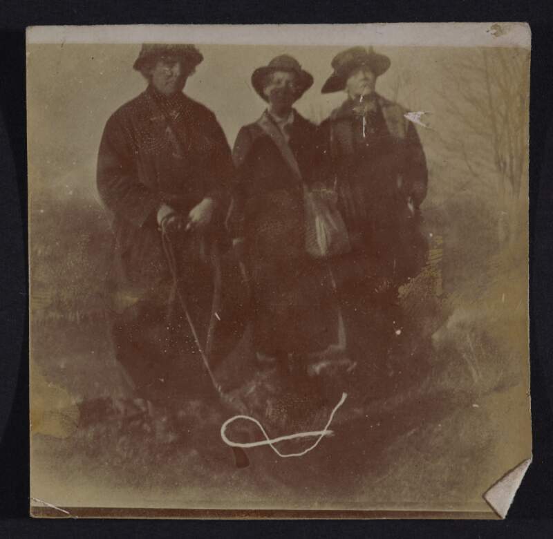 [Three women standing outdoors holding bags, with dog on leash, and tree in background]