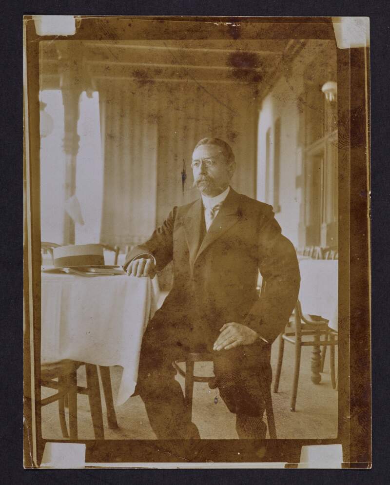 [Portrait of unidentified man, seated, with hat and papers on table]