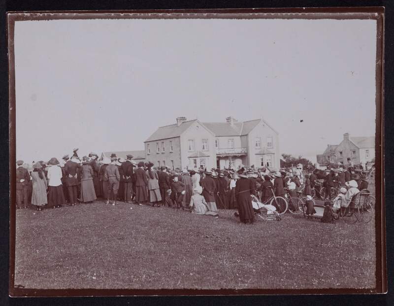 [Crowd of men, women, and children gathered on grassland in front of a large house]