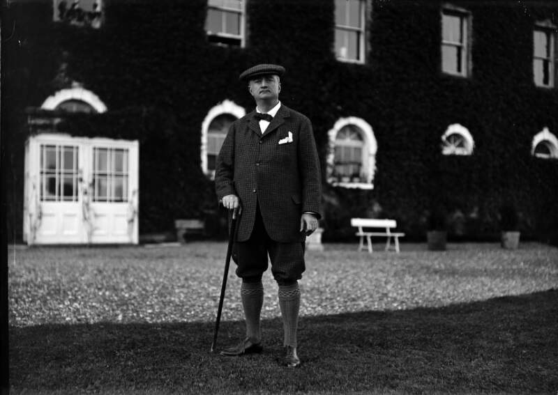 [John Redmond standing on lawn with walking stick, large house in background]