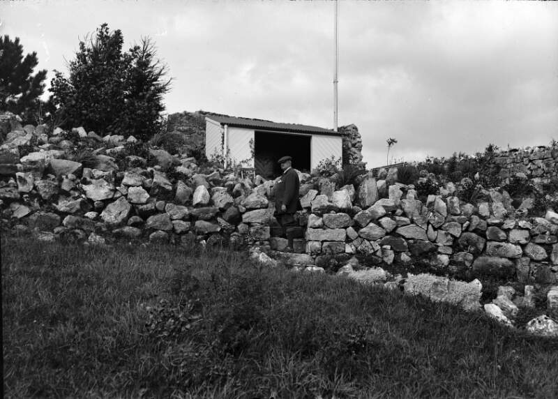 [John Redmond, at stile in stone wall, with shed in background