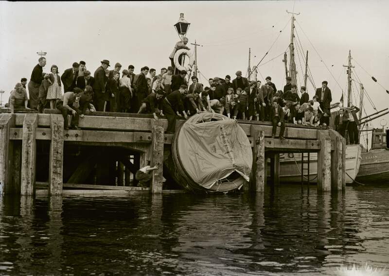 [Crowd on pier watching men drop inflatable raft into ocean, Co. Donegal]