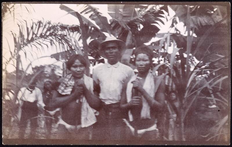[A well-dressed man and two tribespeople, with several others among the trees in the background, set in the Putumayo region]