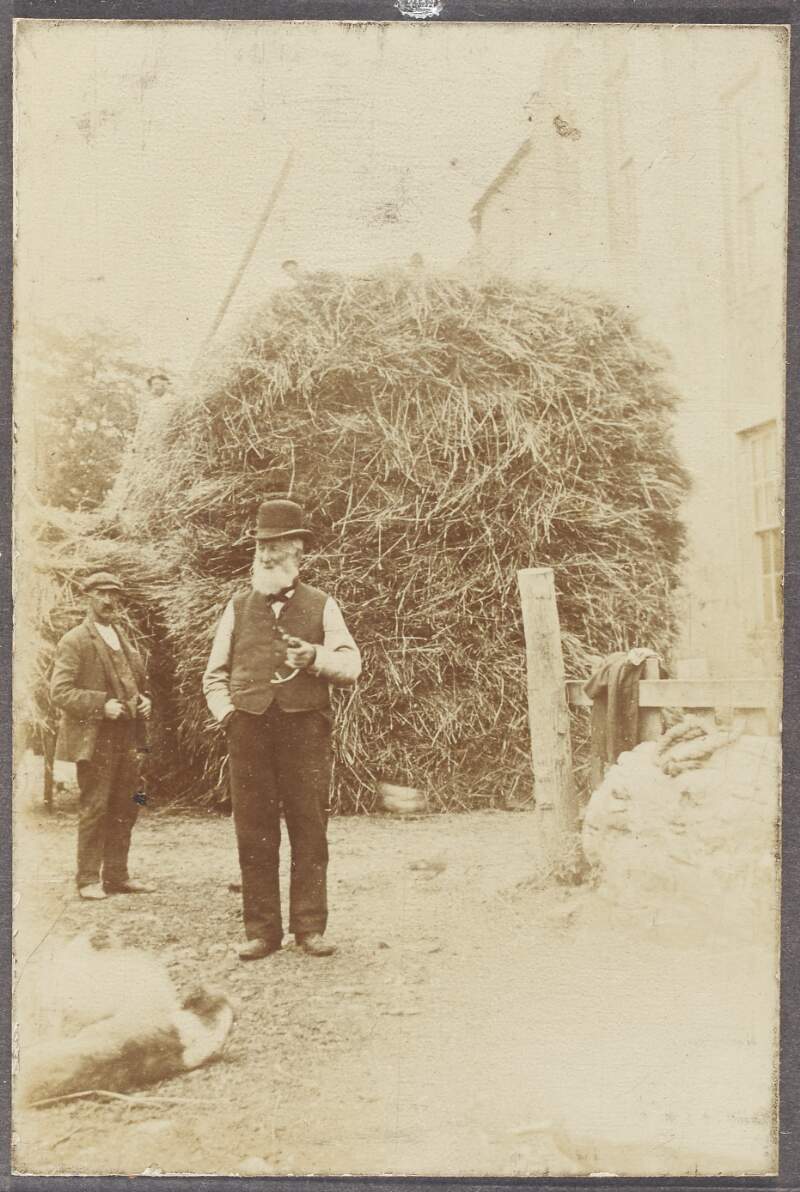 [Photograph of two unidentified men, with a large hay bale in the background]