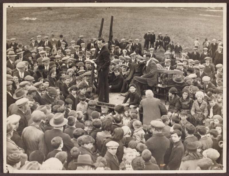 [Man orating on platform, surrounded by a crowd of men, women and children]