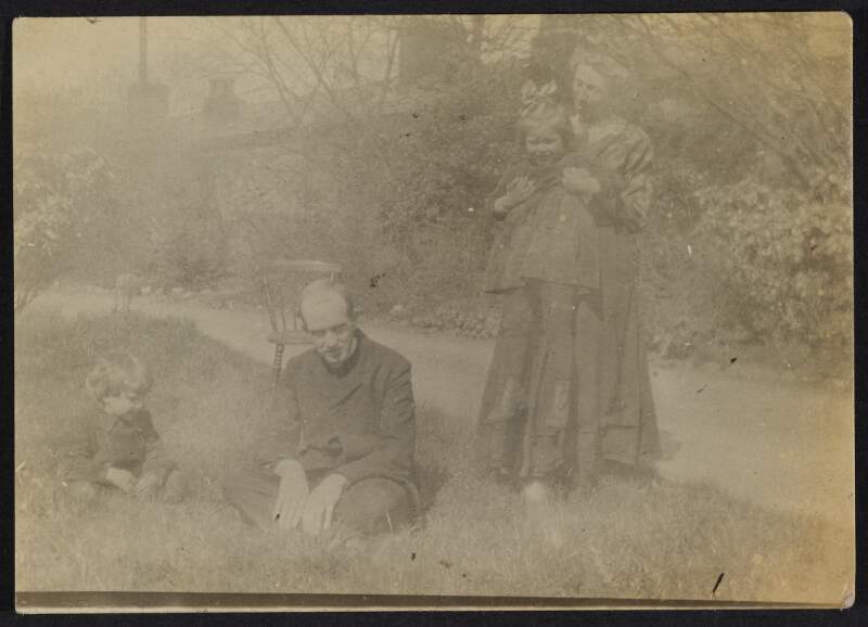 [Unidentified man kneeling on ground with child beside him and unidentified woman standing holding a girl in her arms]