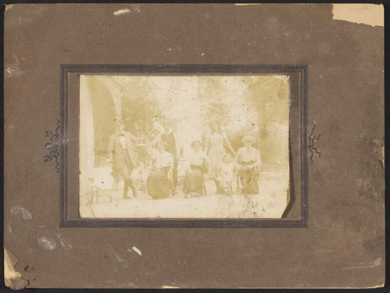 [Group photograph of four women, two men and two young girls posing outside a house]