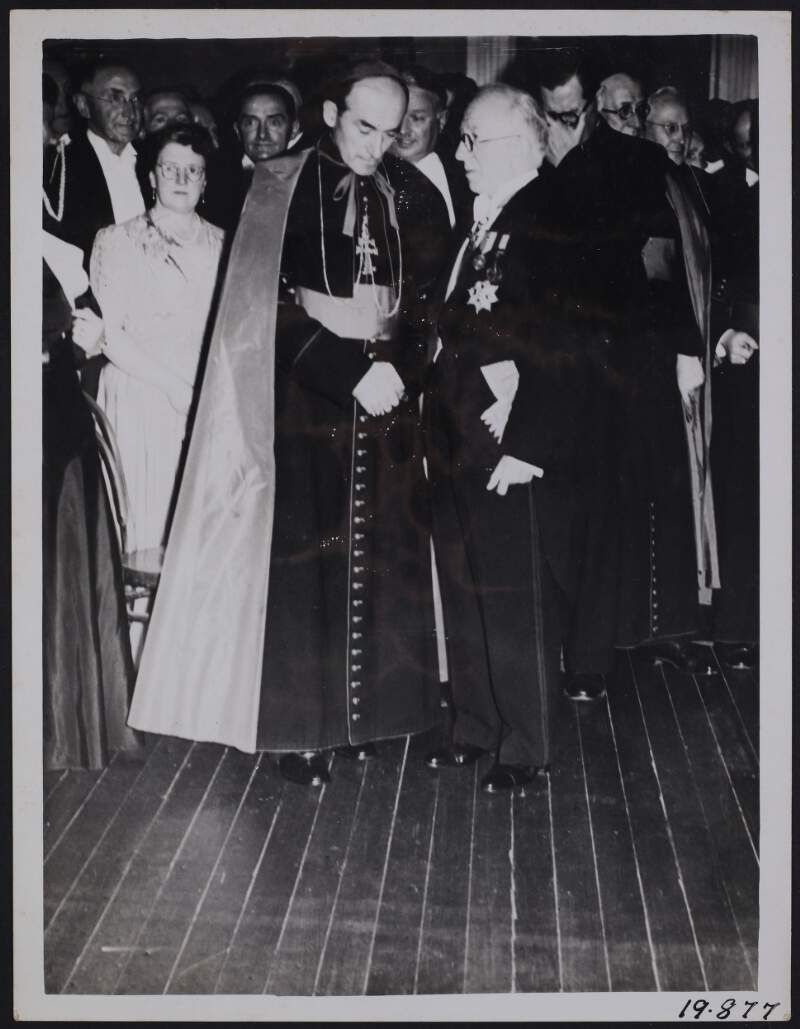 [Seán T. O'Kelly (right) and John Charles McQuaid, Archbishop of Dublin, standing in conversation with a crowd behind them]