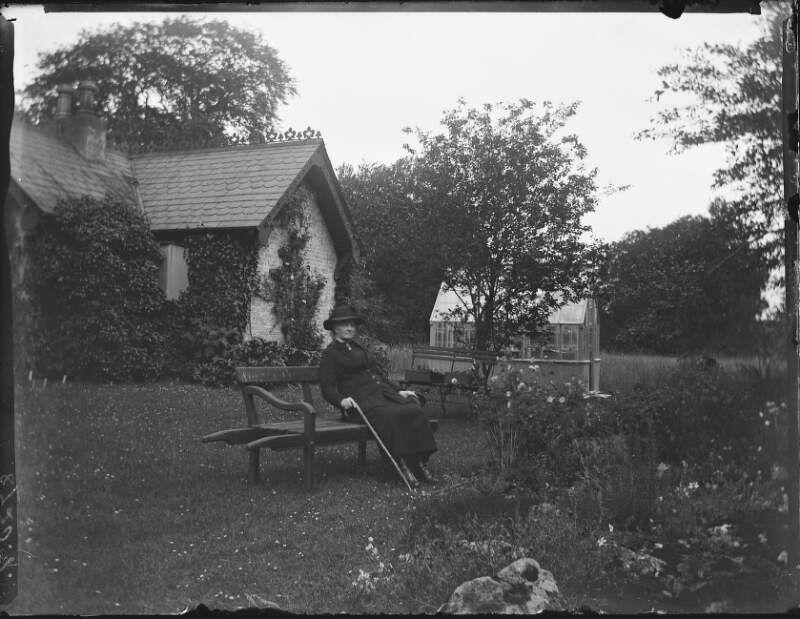 [A.C.C on wheelbarrow seat. Elderly Augusta Dillon on seat in front of photograph house and small glasshouse.]