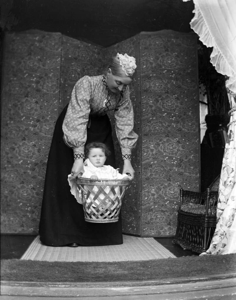 [Augusta Dillon holding toddler in small wicker basket. Chrysanthemum pattern screen in background.]