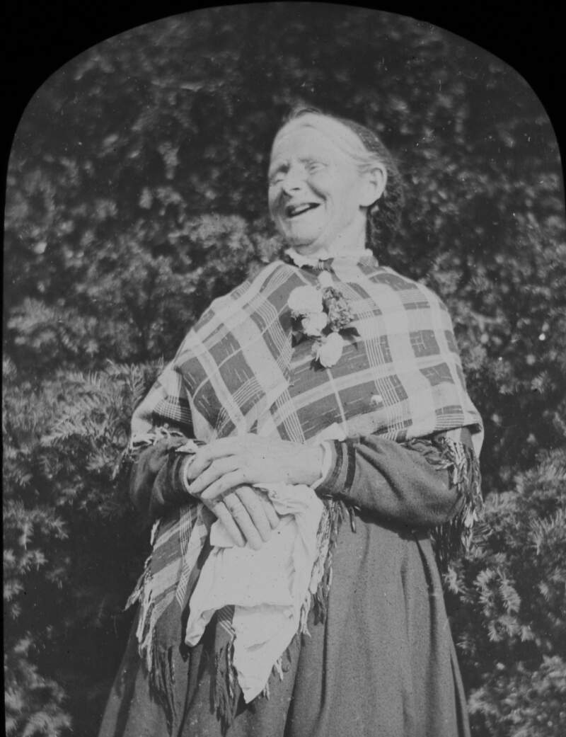[Local woman laughing, wears plaid shawl, long skirt and a rosette.]