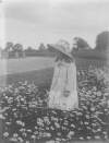 [Little girl in field of flowers. She wears a large bonnet and double breasted jacket, tree in background. Ursula Mahon.]