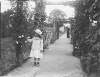 [Ursula Mahon, daughter of Edith Dillon and Sir William Mahon c.1910. Young girl picking roses growing near wooden arches covered in clematis. Pathway surrounded on either side by flower beds, woman in distance holding parasol.]