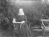 [Augusta Crofton Dillon seated in chair, wears head scarf and veil, bushes in background. Wheel barrow bench visible to left.]