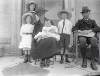 [Ursula on terrace group, 1910. Family group on steps of house includes man, woman and four children, infant on mothers lap, eldest daughter wears pinstripe dress and long hair, two middle children wearing wide brimmed hats and lace up boots.]