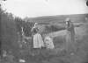 [A stout woman wearing an apron and man wearing a long tweed overcoat and cap with rural landscape in the background.]
