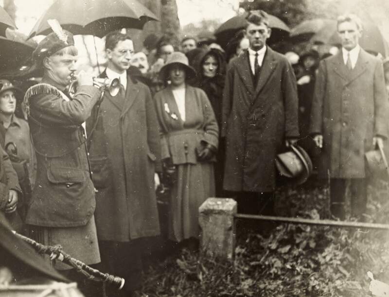 [Éamon De Valera and unidentified men and women standing at an unidentified graveside while a soldier plays the trumpet]