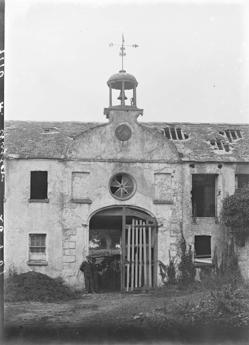[Front of dilapidated building with bell tower, with entrance to courtyard, and also with group of people standing at entrance]